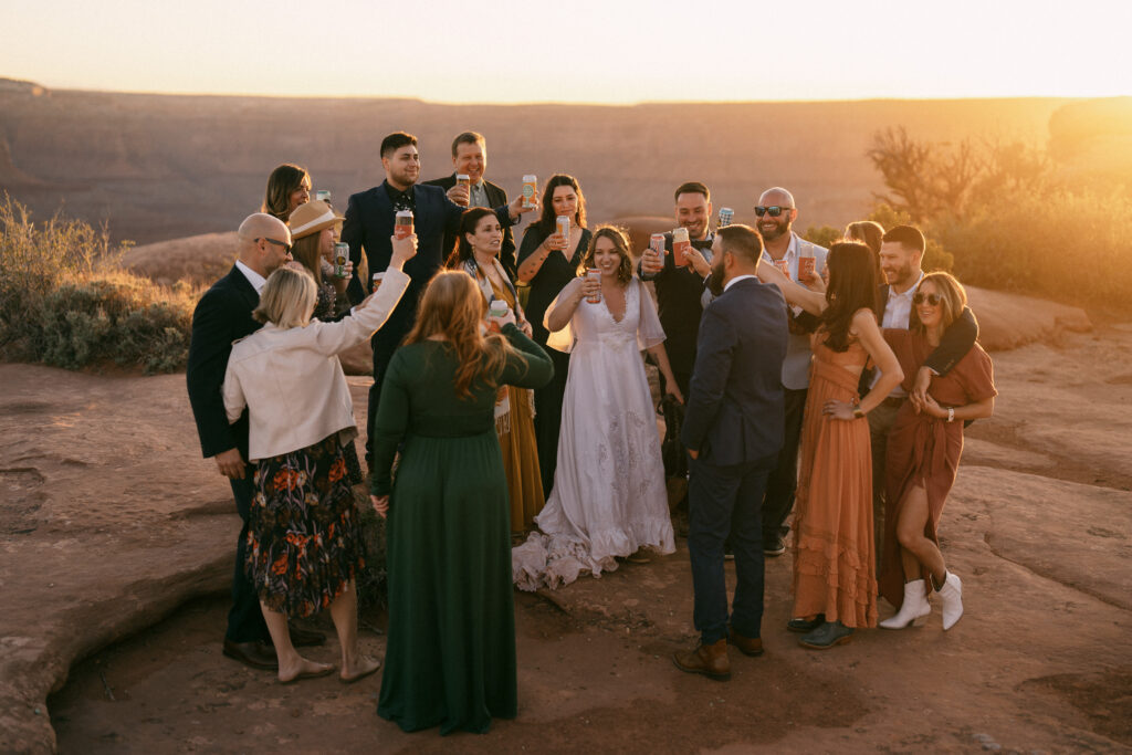 A couple, surrounded by a small group of their family and friends, clinking craft beers following their micro wedding ceremony at sunset in Moab, Utah.