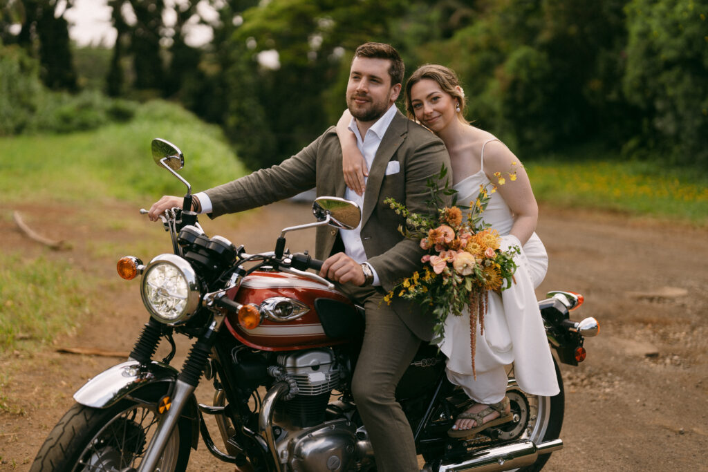 Groom and bride on a vintage motorcycle in their wedding attire, holding their wedding bouquet, in Kauai Hawaii. 