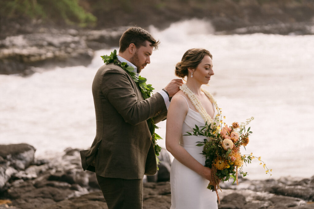 Groom wearing a lei, placing white flower leis around his bride's neck before their elopement ceremony on the beach in Kauai Hawaii. 