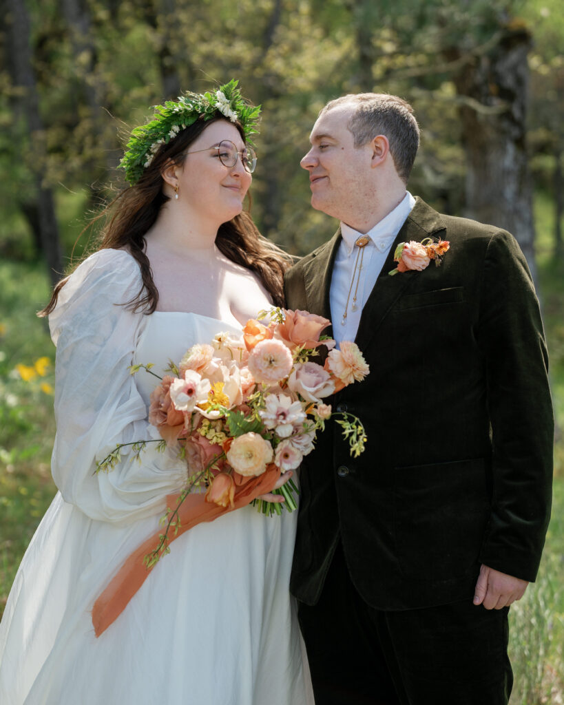 bride and groom portrait on their oregon elopement day, bride wearing a crown of ferns and hold spring blooms in shades of pink and peach